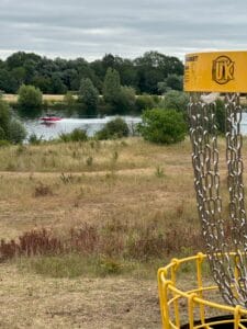 Box End Park Disc Golf – yellow disc golf basket in the foreground, with a water skier and red boat on the lake in the background.